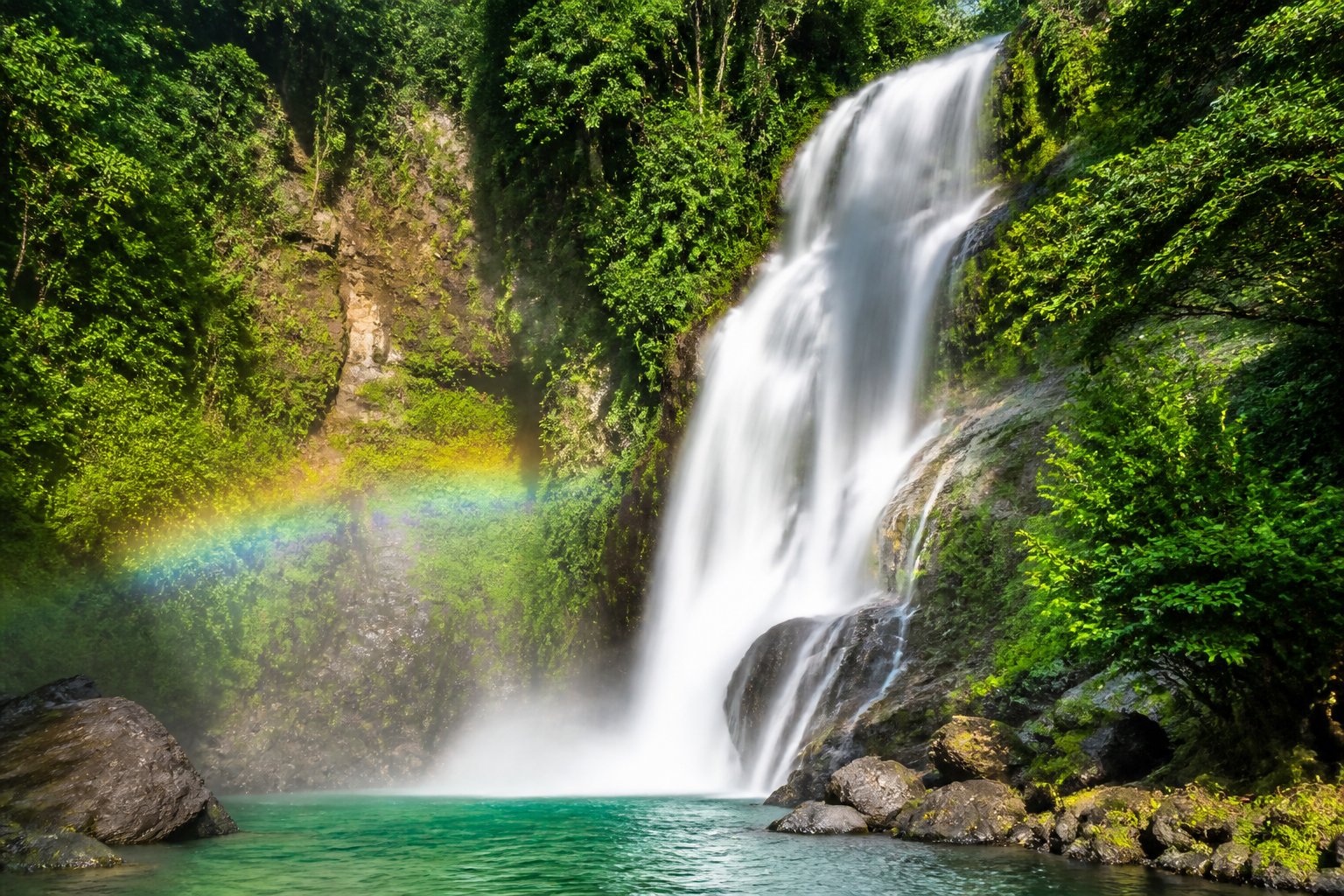Cachoeira majestosa cercada por floresta tropical densa, com água caindo sobre penhascos rochosos e formando uma piscina clara e serena abaixo.