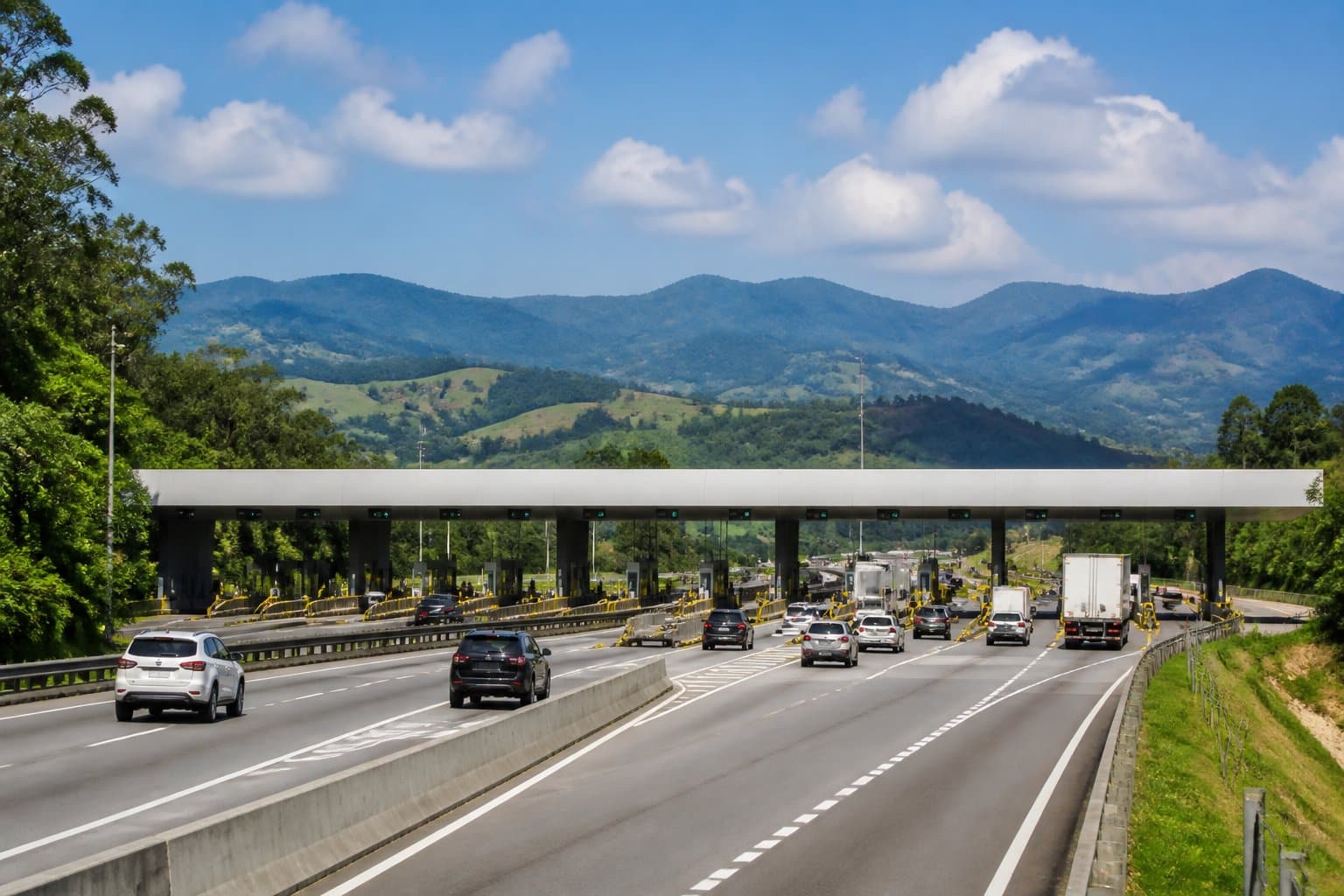 Rodovia com pedágios e veículos passando, cercada por árvores e montanhas ao fundo.